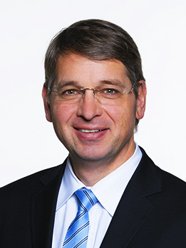 Porträt eines lächelnden Mannes mittleren Alters mit Brille, dunklem Anzug, weißem Hemd und blau gestreifter Krawatte.