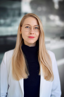 Junge blonde Frau mit Brille, schwarzem Rollkragen und weißem Blazer schaut direkt in die Kamera.