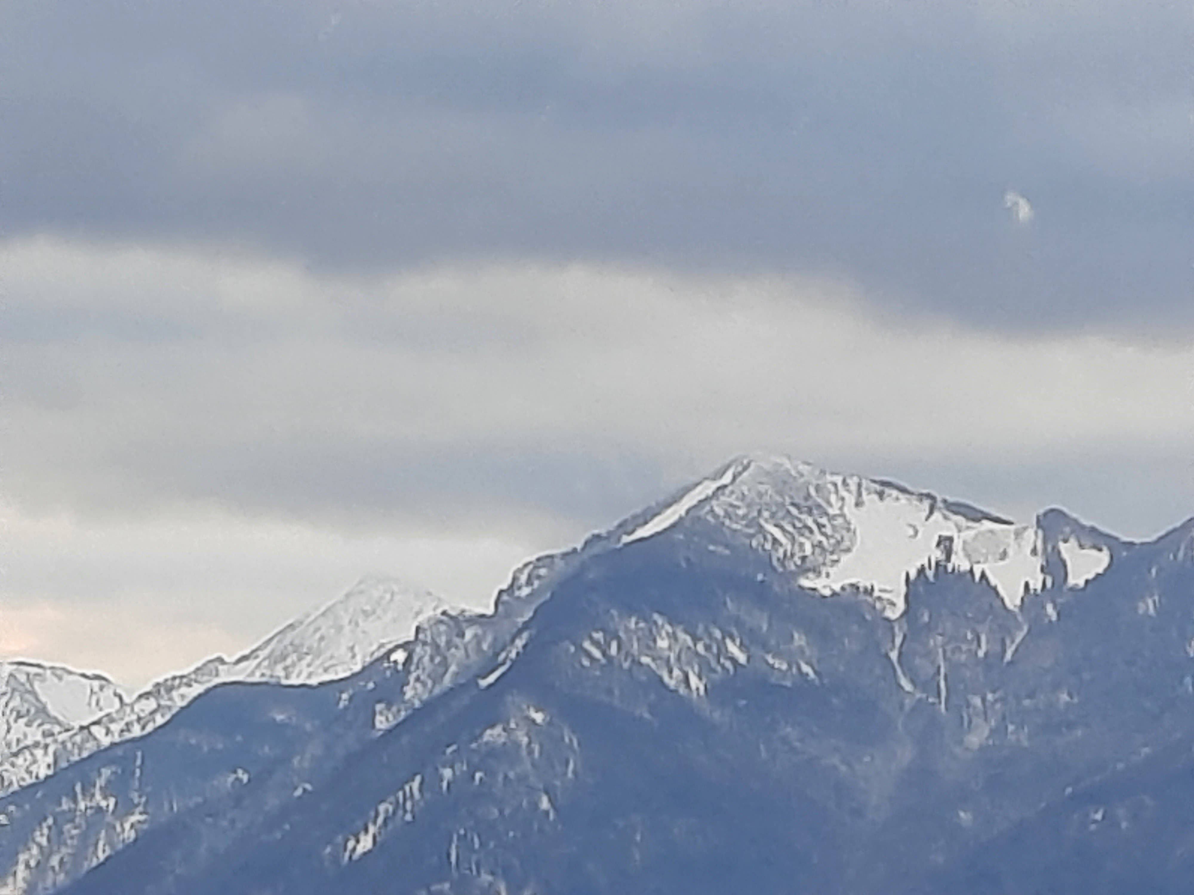 Ferne, schneebedeckte Bergkette unter einem bewölkten Himmel. Dunkle Gipfel sind teilweise mit weißem Schnee bedeckt.