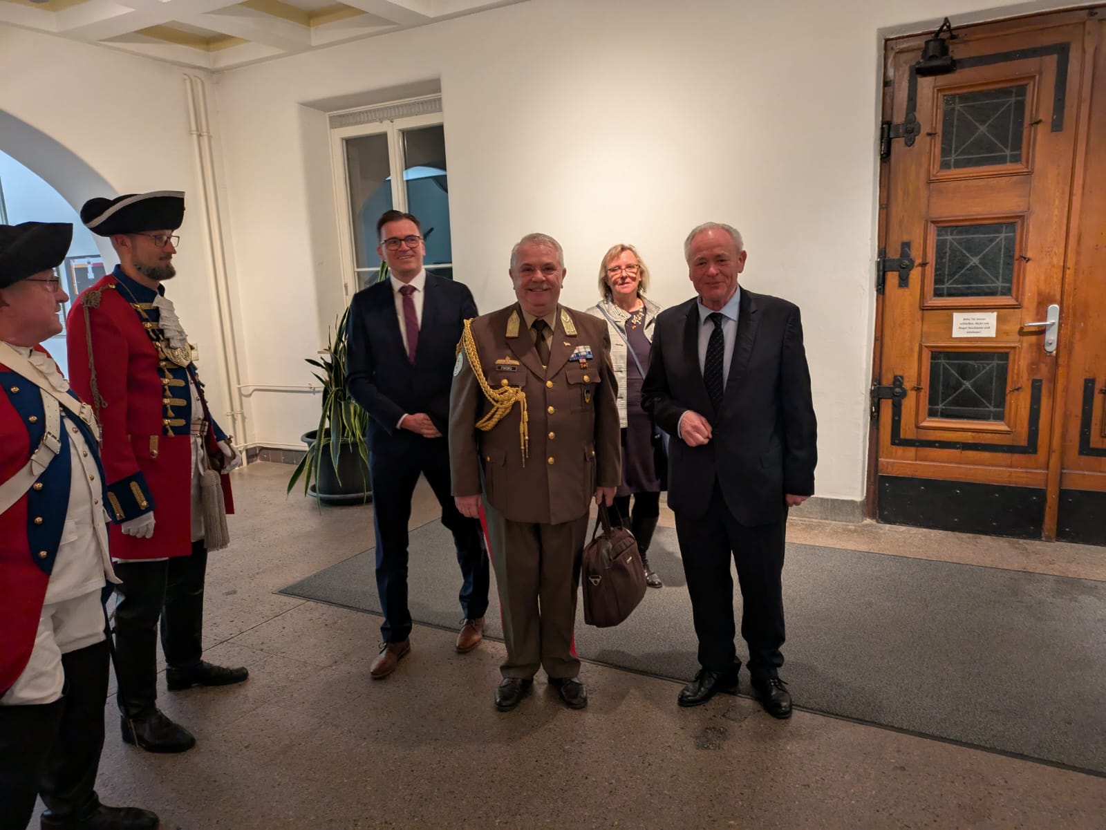 Personen in historischen und modernen Militäruniformen, Anzügen und einer Frau, lächeln drinnen.