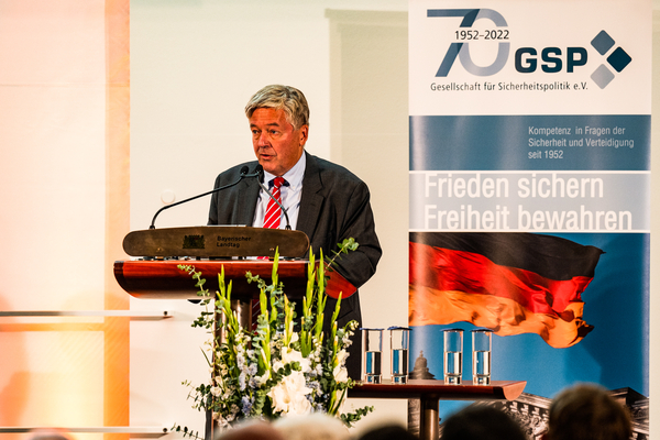 Mann spricht am Rednerpult des Bayerischen Landtags, im Hintergrund ein GSP-Banner zum 70-jährigen Jubiläum mit deutscher Flagge.