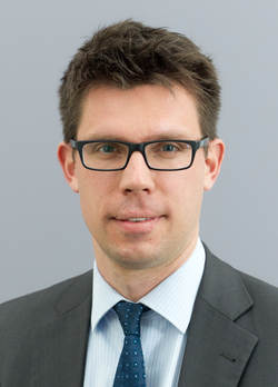 Kopfaufnahme eines professionellen Mannes mit dunklem Haar und Brille, der einen Anzug und eine gemusterte Krawatte trägt.