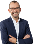 Ein lächelnder Mann mit Brille, in einem dunkelblauen Anzug und weißem Hemd, die Arme verschränkt vor weißem Hintergrund.