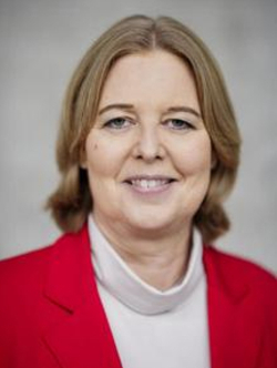 Porträtaufnahme einer lächelnden Frau mit blonden Haaren, die einen roten Blazer und ein helles Rollkragenoberteil trägt.