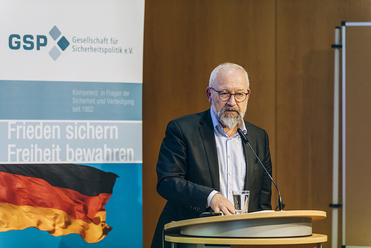 Bärtiger Mann mit Brille spricht am Rednerpult vor einem GSP-Banner, das die deutsche Flagge zeigt.