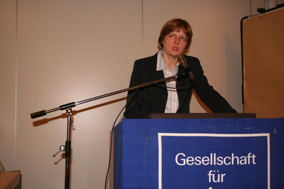 Frau in gestreiftem Blazer spricht am Mikrofon hinter einem Rednerpult mit "Gesellschaft für" Aufschrift auf blauem Tuch.