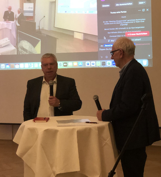 Zwei Männer mit Mikrofonen stehen an einem Tisch, einander zugewandt, vor einer großen Leinwand mit einer Online-Meeting-Chat-Oberfläche.