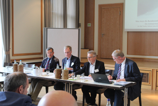 Vier Männer an einem Konferenztisch während einer Podiumsdiskussion, mit einer Projektionswand im Hintergrund.