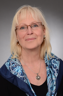 Kopfportrait einer lächelnden blonden Frau mit Brille, blau gemustertem Schal und Perlenkette.