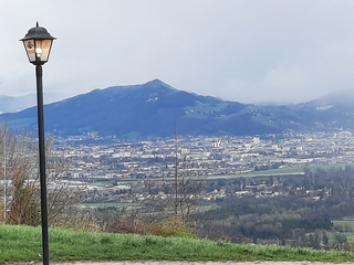 Ein Laternenpfahl im Vordergrund, der eine Stadt in einem Tal mit Bergen unter bewölktem Himmel überblickt.