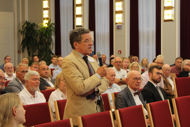 Ein Mann in beigem Sakko und Brille spricht in ein Mikrofon, umgeben von einem Publikum in einem Saal.