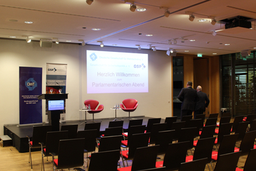 Konferenzraum, Bildschirm zeigt 'Herzlich Willkommen zum Parlamentarischen Abend', daneben Logos und Podium. Viele Stühle, zwei Männer verlassen den Bereich.