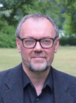 Kopfaufnahme eines Mannes mit grauen Haaren, Bart und schwarzer Brille, der ein dunkles Hemd und Sakko trägt, im Freien.