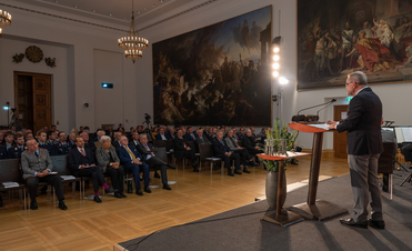 Vortrag in einem großen Saal mit Publikum, Redner am Pult und beeindruckenden Wandgemälden.