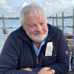 Ein lächelnder Mann mit grauen Haaren und Bart sitzt an einem Tisch am Wasser, eine Serviette in seiner Tasche.