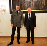 Zwei Männer, einer in Militäruniform und einer im dunklen Anzug, stehen lächelnd nebeneinander in einem Raum.