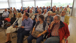 Zuhörer, hauptsächlich Senioren, lauschen aufmerksam einer Veranstaltung in einem gut beleuchteten Raum mit großen Fenstern.