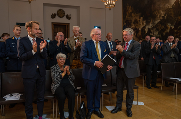 Eine Gruppe von applaudierenden Menschen, darunter Männer in Anzügen und eine Frau, in einem festlichen Saal.