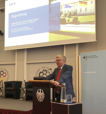 Mann spricht am Rednerpult mit Bundesadler, vor Leinwand mit "Begrüßung", Namen und Branding der Bundesakademie.