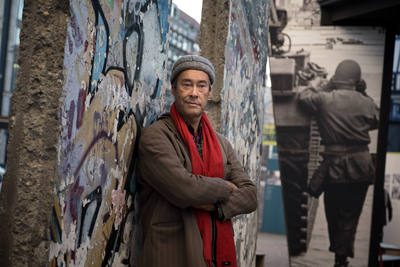 Mann mit rotem Schal und Mütze lehnt an einer graffitibedeckten Mauer, mit verschwommenem historischem Foto im Hintergrund.