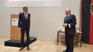 Zwei Männer in Anzügen in einem Konferenzraum mit deutschem Adlerpult und Flagge. Ein Mann trägt eine Maske am Handgelenk.