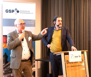 Zwei Männer bei einer GSP-Veranstaltung: Ein älterer Redner gestikuliert mit Mikrofon, ein jüngerer Mann mit Bart und Headset steht am Pult.