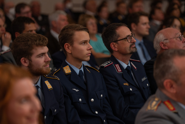Drei Männer in deutschen Militäruniformen, darunter ein junger Luftwaffe-Angehöriger, sitzen aufmerksam im Publikum.