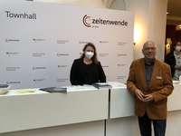 Zwei Personen, eine maskierte Frau und ein Mann im braunen Sakko, stehen an einem Tresen vor einer 'Townhall zeitenwende on tour'-Kulisse.