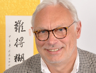 Porträt eines lächelnden Mannes mit grauen Haaren und Brille vor einem Hintergrund mit asiatischer Kalligrafie.