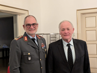 Zwei Männer, einer in Militäruniform mit Medaillen und Brille, der andere in schwarzem Anzug, stehen lächelnd in einem Raum.