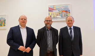 Drei Männer in Anzügen, einer bärtig mit Brille, posieren vor einer weißen Wand mit zwei farbigen Stadtansichts-Gemälden.