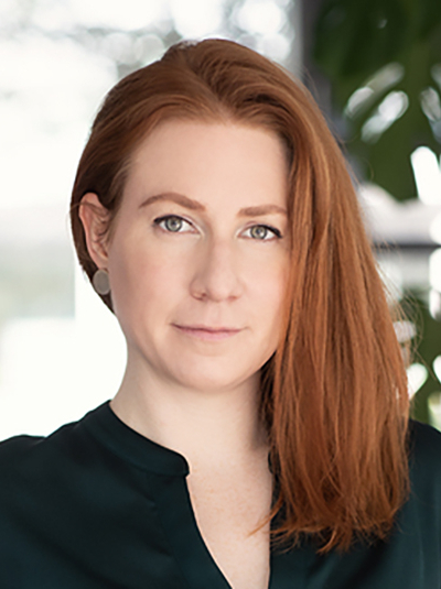 Porträt einer Frau mit langen roten Haaren, hellen Augen und heller Haut, die ein dunkelgrünes Hemd trägt und direkt in die Kamera blickt.