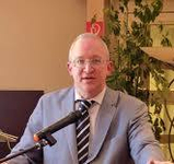 Ein professioneller Mann im Anzug und mit Brille spricht in ein Mikrofon bei einer Präsentation.