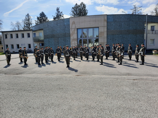 Eine Militärkapelle in dunkelgrünen Uniformen und Baretten steht mit Instrumenten vor Gebäuden unter klarem Himmel.