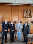Vier Personen, drei Männer und eine Frau, stehen in einem offiziellen Raum mit Holzwänden, deutscher Flagge und Bundesadler.