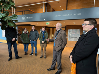 Eine Gruppe Menschen hört einem Mann in der UN Bonn Lobby zu, mit einem Schild 'Shaping a Sustainable Future' darüber.