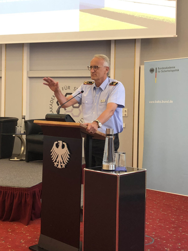 Mann in Uniform spricht an einem Rednerpult mit deutschem Adleremblem. Banner der Bundesakademie für Sicherheitspolitik im Hintergrund.