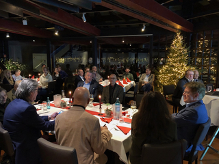 Menschen versammeln sich zu einem festlichen Abendessen in einem gedämpft beleuchteten Raum mit Tischen, Kerzen und einem leuchtenden Weihnachtsbaum.