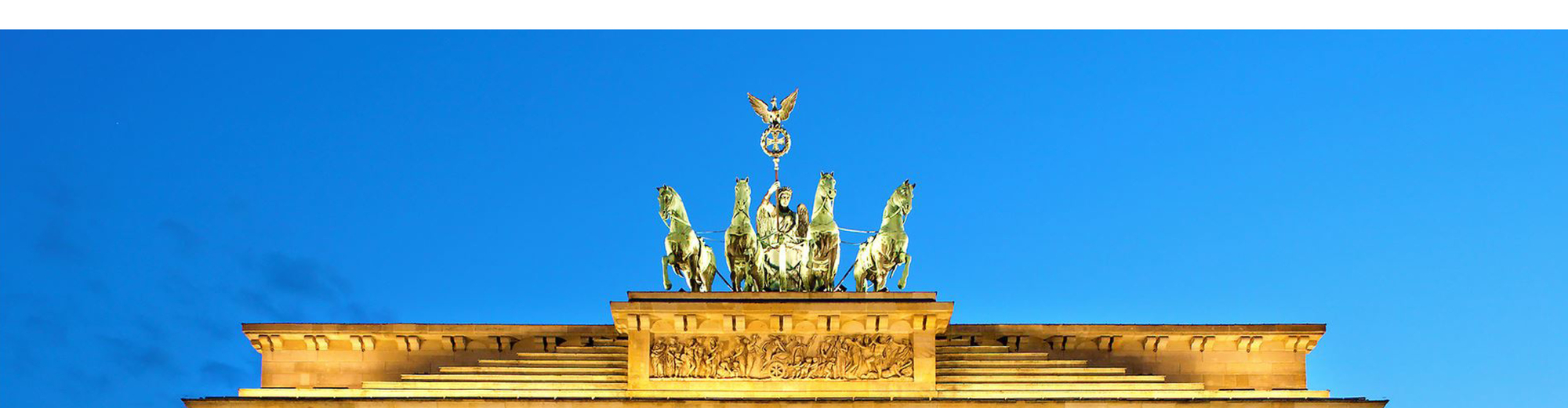Die Quadriga-Statue mit Victoria und vier Pferden auf dem Brandenburger Tor, beleuchtet vor blauem Himmel.