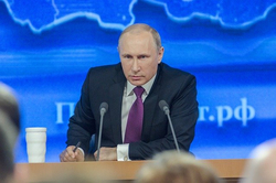 Wladimir Putin sitzt an einem Pult mit Mikrofonen, hält einen Stift und blickt ernst in die Kamera bei einer Pressekonferenz.
