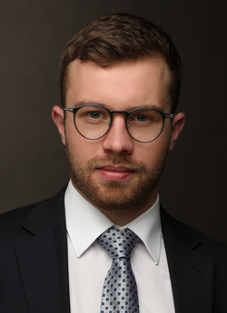 Ein professioneller Mann im dunklen Anzug, weißem Hemd, gemusterter Krawatte und Brille, blickt direkt in die Kamera.