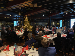 Abendveranstaltung in einem Restaurant mit vielen Gästen, festlich beleuchtetem Weihnachtsbaum und Präsentationsbildschirmen.