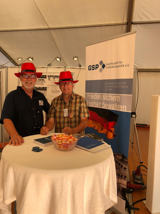 Zwei lächelnde Männer mit roten Hüten und Brillen stehen an einem GSP-Stand mit deutscher Flagge, Broschüren und Süßigkeiten.