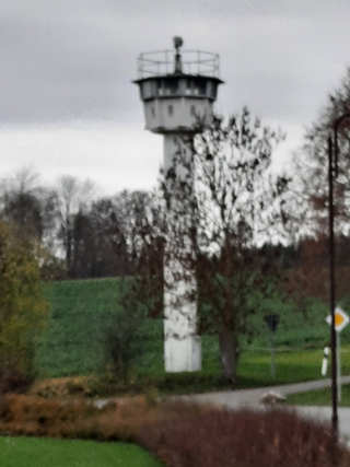 Verschwommener hoher Beton-Wachturm mit runder Aussichtsplattform, umgeben von grünen Feldern und Bäumen unter bewölktem Himmel.