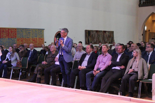 Mann spricht mit Mikrofon vor sitzendem Publikum bei einer Indoor-Veranstaltung.