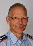 Kopfaufnahme eines Mannes mit Brille und Schnurrbart in hellblauem Uniformhemd mit Rangabzeichen auf den Schultern.