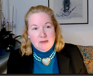Eine blonde Frau in einem blauen Rollkragenpullover, schwarzem Blazer und türkiser Halskette mit Muschelanhänger blickt leicht weg.