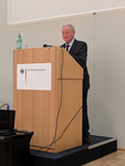 Älterer Mann im dunklen Anzug steht hinter einem Holzpult mit "Bundessprachenamt"-Schild, Mikrofon und Wasser.