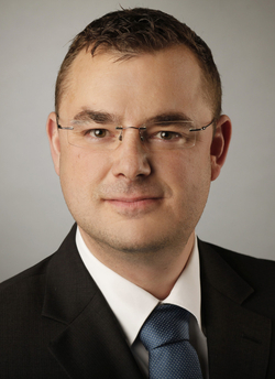 Porträt eines Mannes mit kurzem braunem Haar und Brille, in dunklem Anzug und blauer Krawatte, blickt in die Kamera.