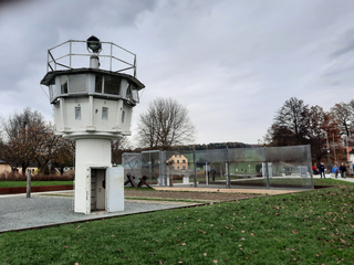 Ein weißer ehemaliger Grenzturm mit Scheinwerfer, Zaunabschnitt, kahlen Bäumen und Rasen mit Herbstlaub unter bewölktem Himmel.